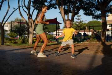 Fototapeta premium Smiling athletic mother and her energetic young son running playfully together in a sunny park, enjoying quality family time, movement, and fitness in a joyful and active outdoor setting