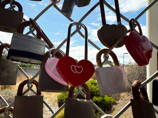 Multiple padlocks including red heart lock hanging on wire fence with natural background and clouds, expressing concept of love, memory, promise, and human emotion