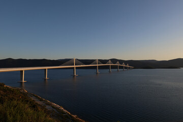 Obraz premium Scenic view of the modern Peljesac Bridge in Croatia spanning the calm Adriatic Sea during golden hour against dark mountains.