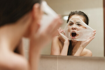 Surprised Asian woman applying a facial mask in front of a bathroom mirror, showcasing self-care and beauty routine with natural light.