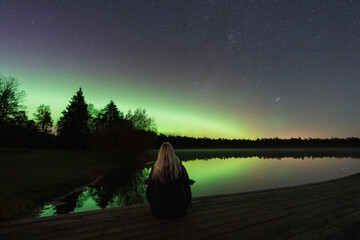 A woman peacefully watching the magical Northern Lights (Aurora Borealis) and starry sky reflected in a serene Estonian lake.