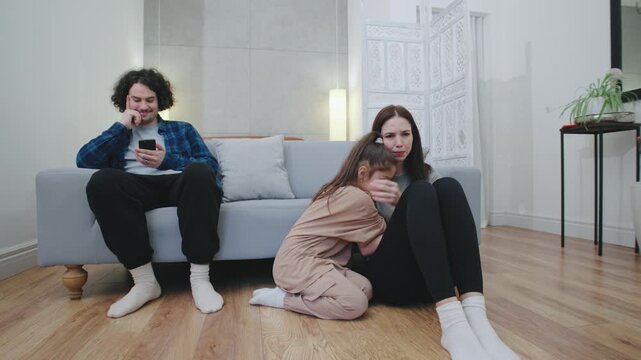 A teenage girl gently comforts her crying mother on the floor, while the father sits on the sofa, smiling at his phone, detached from the emotional scene unfolding