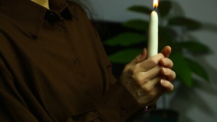 Woman hand holding burning white candle in room interior background with blur green flower. Candle fire flame with space. Magic flaming of one candle fire