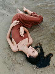 A relaxed female model lies on damp sand wearing a rust red dress, creating a striking silhouette against the beach surface with soft light, subtle shadows, and an elegant pose.