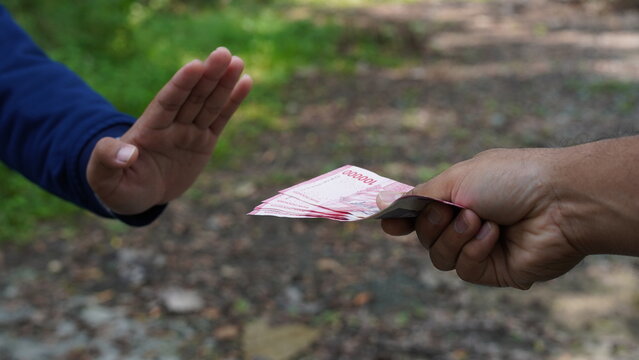 One hand offers Indonesian rupiah while another hand firmly rejects the money with a clear "stop" gesture