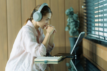 Smiling Asian female student wearing headphones for online learning using AI, listening to lectures, watching webinars, writing notes, and sitting in a coffee shop with a tablet.