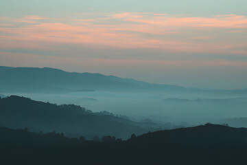 Mountain Serenity: The tranquil mountain scape unfolds with layers of rolling hills draped in a soft, ethereal mist, kissed by the gentle blush of the sunrise. An invitation to calm, peace.