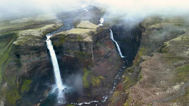Haifoss waterfall emerges dramatically from steep cliffs in Iceland, surrounded by lush greenery. Mist rises, creating an enchanting atmosphere at dusk. A true natural wonder for adventure seekers.