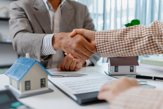 Two people in business attire finalize a real estate deal, shaking hands across a desk with house models, keys, and a contract, symbolizing agreement, trust, and successful negotiation.