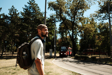 Young man with a backpack standing near tram tracks in a park on a sunny day. Concept of travel, transport, and urban lifestyle