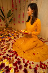 Happy young Indian woman wearing traditional yellow clothes holding plate with oil lamps and flowers celebrating Diwali festival. Selective focus. Festival of lights. Deepawali.