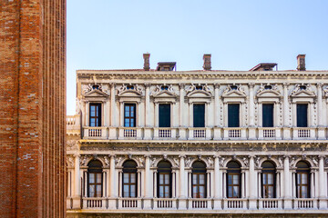 The Renaissance-style facade of the Procuratie Vecchie beside the brick Campanile di San Marco in Piazza San Marco, Venice, under a clear blue sky. 