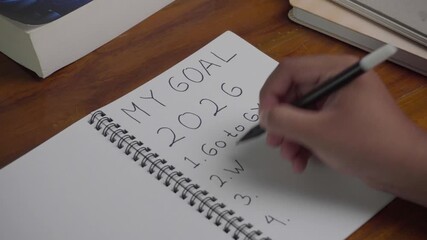 Person wrote "Wake up Early" in his 2026 goal on his notebook. On a wooden table.