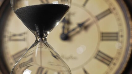 Close up of an hourglass with black sand flowing symbolizing the passage of time urgency and deadlines set against a blurred vintage clock face in the background.