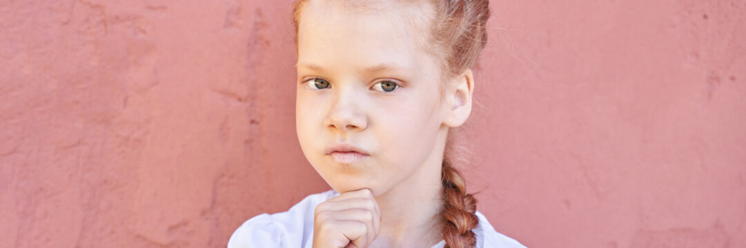 Thoughtful caucasian female child with red hair against pink wall in white shirt.