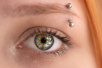 Naklejka premium Close-up portrait of a young woman with striking blue eyes and orange hair, showcasing unique facial piercings and natural beauty in a soft, blurred background