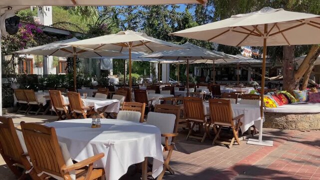 An elegant outdoor restaurant terrace features neat rows of wooden tables with white cloths and chairs, shaded by large umbrellas on a sunny day, perfect for dining and relaxation.