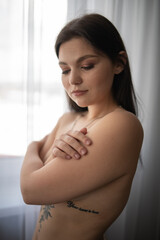 Young woman with long dark hair poses gracefully, embracing herself in soft natural light,...