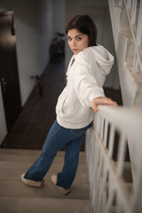 Young woman with short hair wearing a white hoodie leans against a wall in a modern indoor setting, showcasing a casual and relaxed atmosphere with stylish attire