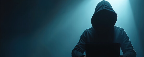 Hacker sits behind laptop in dark hood. Man hacks system. Cybercrime occurs on blue neon background. Threat from network criminal causes info security breach.