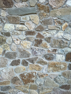 A detailed, full-frame shot showcasing the rugged, irregular texture of a stone wall constructed with various sizes of natural, multi-toned fieldstones and visible mortar joints.
