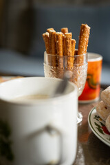Savory breadsticks in a glass between mugs with drinks on a table as refreshments.
