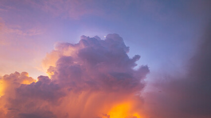 A vivid and dynamic sky captures towering storm clouds illuminated by the warm hues of a setting sun, creating a striking contrast between light and shadow above a darkened forested landscape.