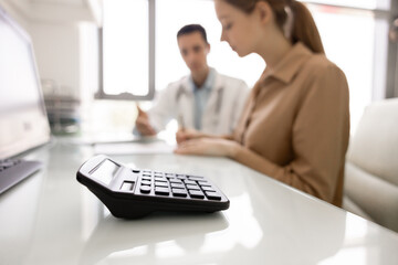 Calculator on desk, blurred doctor and patient discussing healthcare costs