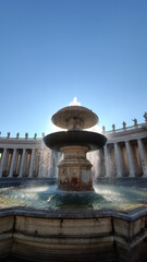 Majestic stone fountain with cascading water under bright sunlight and clear blue sky. Classical architectural design with colonnade and statues in the background, representing European heritage