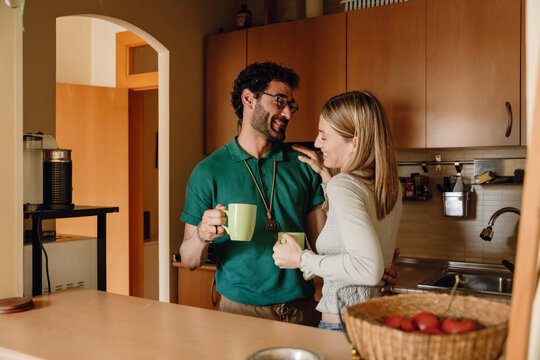 A man hugs a woman who has put her hand on his shoulder while they stand and hold cups while smiling