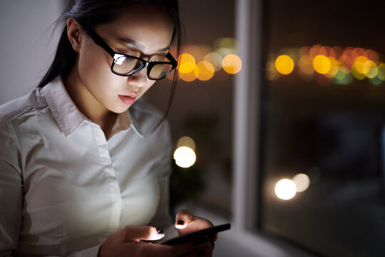 Young Asian female corporate worker texting on smartphone while standing by office window in the late evening, blurred lights in the background