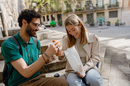 A woman holds a bag and gives a pastry to a man sitting next to her on the stairs while they laugh