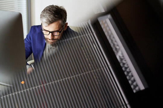Middle-aged male Caucasian manager in glasses working on computer in office, venetian blind seen in the foreground