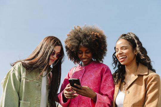 Two female friends are smiling and looking at the phone one of them is holding while a female friend is standing next to them and laughing