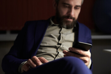 Portrait of defocused Caucasian bearded businessman reading economic news on his smartphone while sitting in office in the late evening