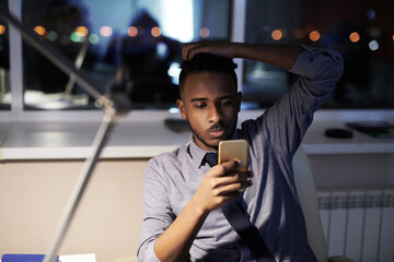 Young African American office manager resting in the end of working day, sitting at table and checking social media on modern smartphone