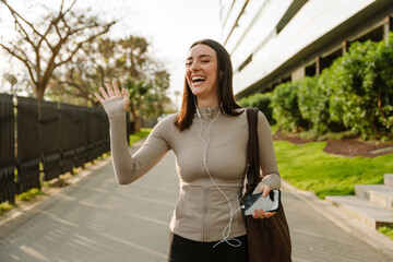 Fototapeta premium Female athlete waving and laughing while holding phone and listening in headphones