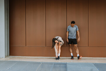 Male and female athletes stand and lean on a wall while she reaches for her feet with her hand