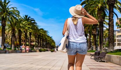 Fototapeta premium Beautiful mature woman tourist walking on seaside boulevard on summer day. Back view