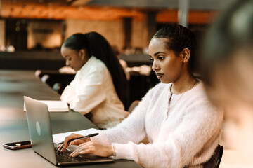 A female student is holding a pen and typing on a laptop while sitting at a table between two female students