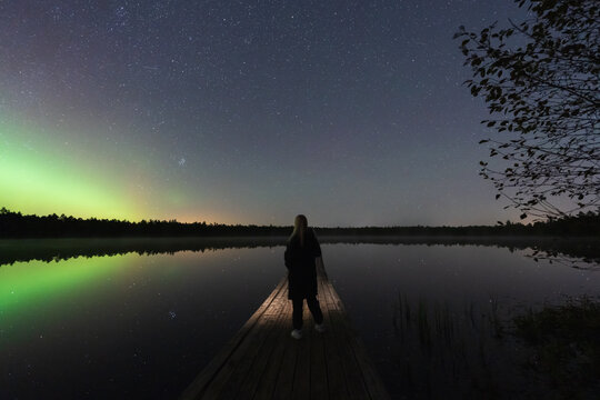 Awe-inspired person on a wooden pier watching the magical Northern Lights, Aurora Borealis, reflect in a calm Estonian lake.