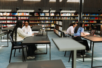 A group of four female students sit at tables while two of them talk and two look at a laptop