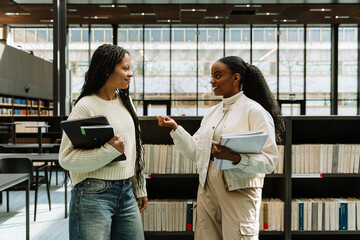 Two female students smiling and holding notebooks and laptop while standing near bookshelves and talking