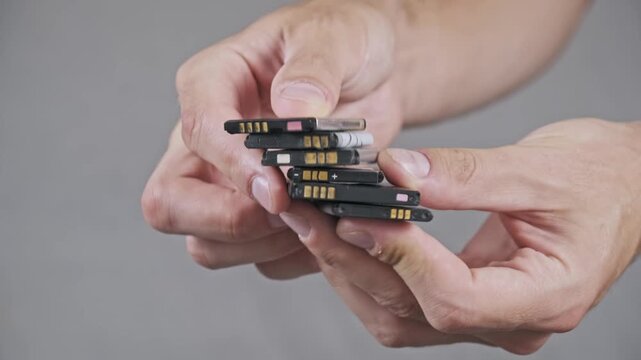 Close-up of hands holding a stack of old used lithium-ion batteries from mobile phones. E-waste recycling and hazardous material disposal theme against a gray background.