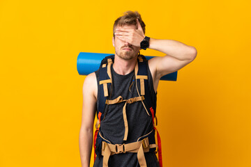 Young mountaineer man with a big backpack isolated on yellow background covering eyes by hands. Do...