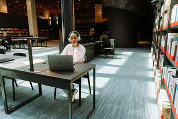 A female student is concentrating on a laptop and sitting at a table while listening in headphones