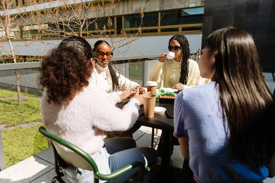 A group of four female students sit at a table while three of them listen to another and one of them drinks from a cup