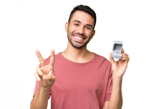 Young handsome caucasian man holding a engagement ring over isolated background smiling and showing victory sign