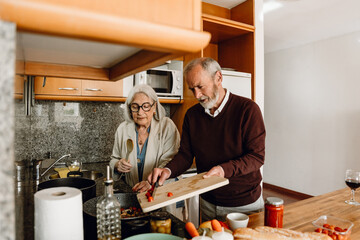 A man pours tomatoes from a cutting board into a frying pan with a knife while a woman stands next to him holding a spoon