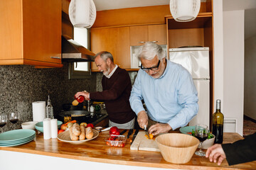 A man is chopping vegetables while a male friend stands next to him, pouring sauce from a jar into a pan